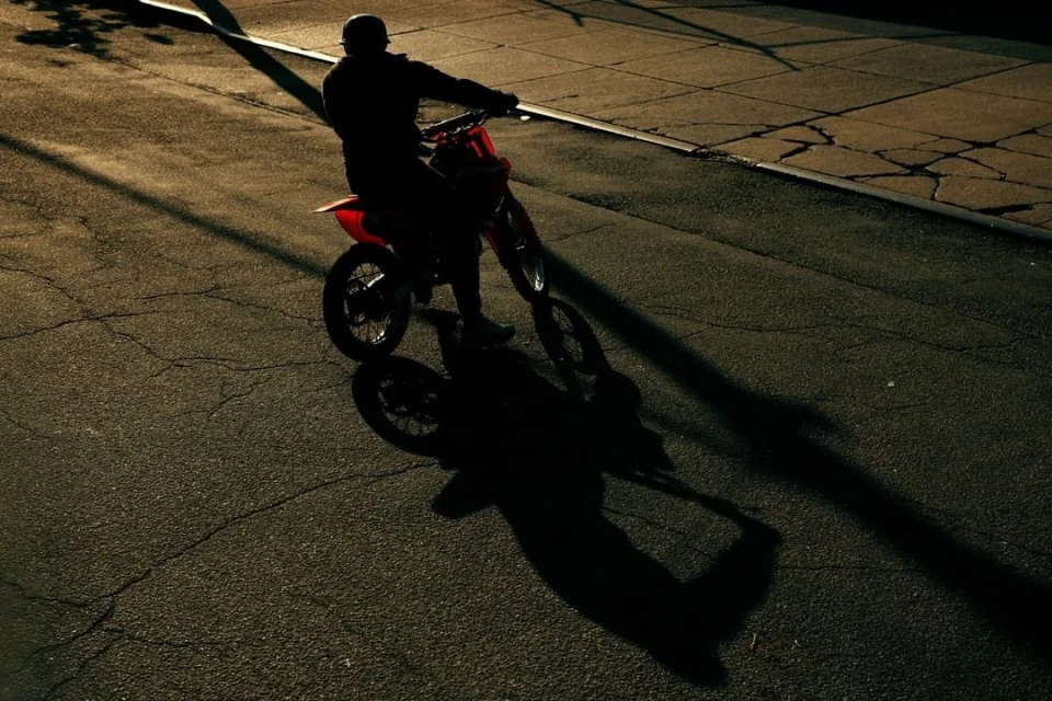A person rides a red dirt bike on an asphalt road at dusk, creating a silhouette and long shadow. The scene is moody and urban.