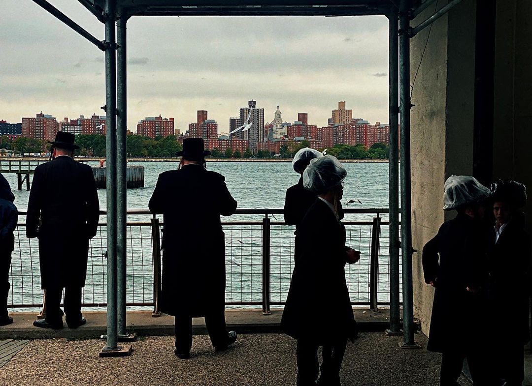 Orthodox Jewish men in traditional attire observe the waterfront of an urban cityscape from an outdoor observation point on an overcast day.