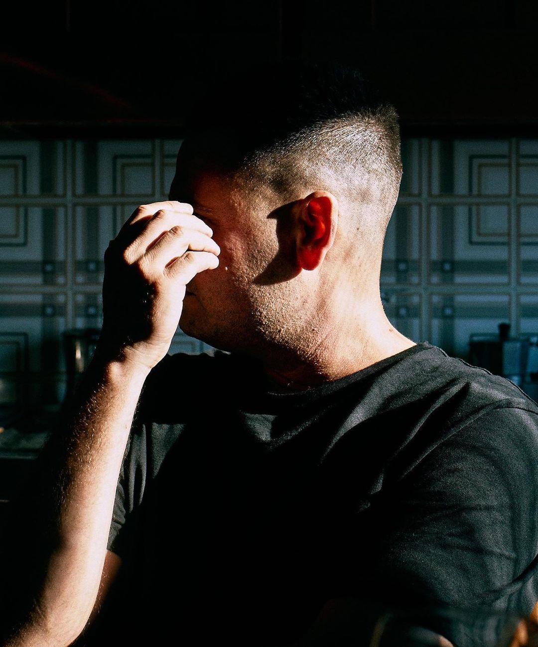 Close-up of a contemplative adult male partially covering his face with his hand, set against a tiled indoor background.