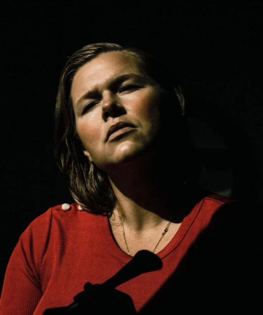 Portrait of adult woman with fair skin and shoulder-length blonde hair wearing a red top in dark, shadowed photo studio setting.