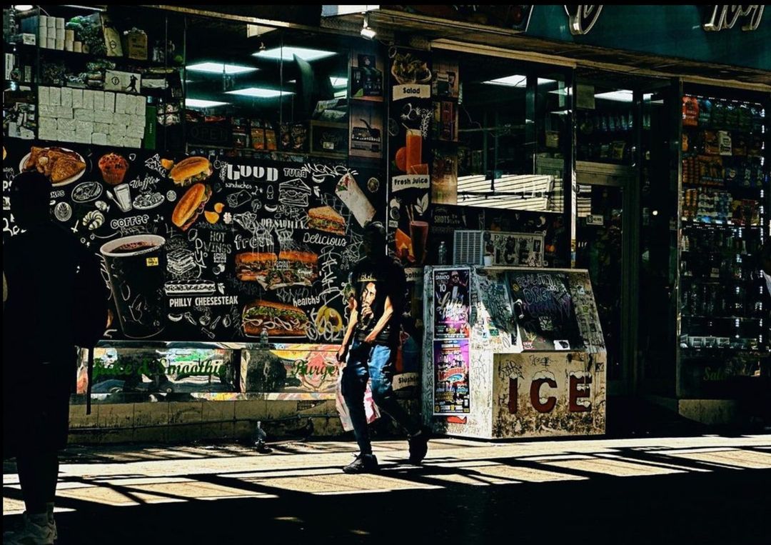 Adult on urban sidewalk walks past food mural on building; an ice box is on the street; natural light.