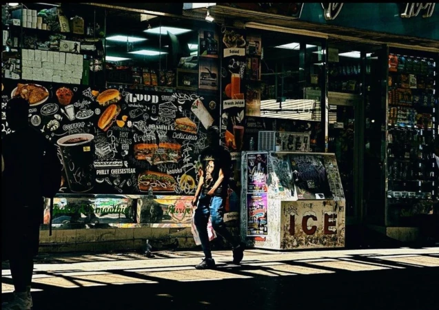 Adult on urban sidewalk walks past food mural on building; an ice box is on the street; natural light.
