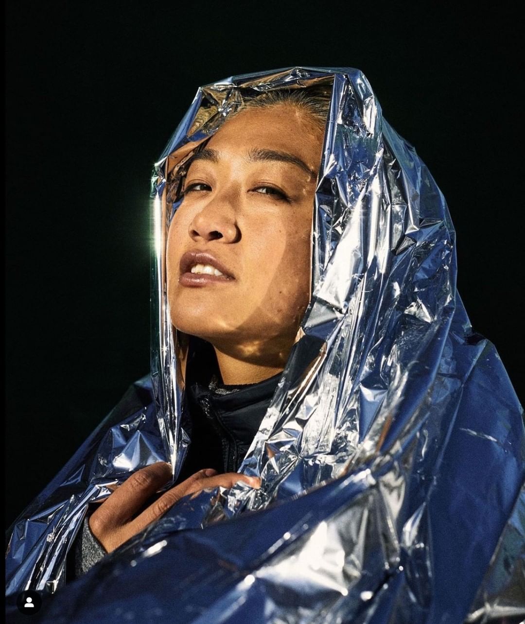 Close-up of an adult female with light skin and wet hair, wrapped in a silver metallic blanket against a stark black background, looking upwards.