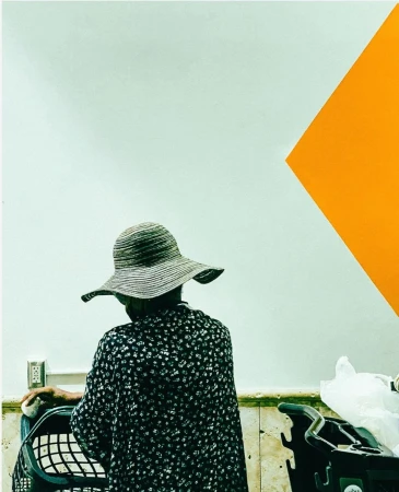 Back view of an older woman wearing a hat pushing a shopping cart indoors against a white wall with an orange geometric shape.