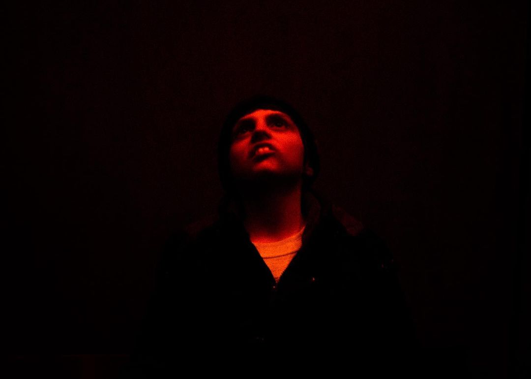 A man with dark hair looks upward in a dark room, illuminated by red lighting, wearing a dark jacket with a lighter shirt underneath.