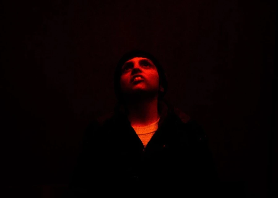 A man with dark hair looks upward in a dark room, illuminated by red lighting, wearing a dark jacket with a lighter shirt underneath.
