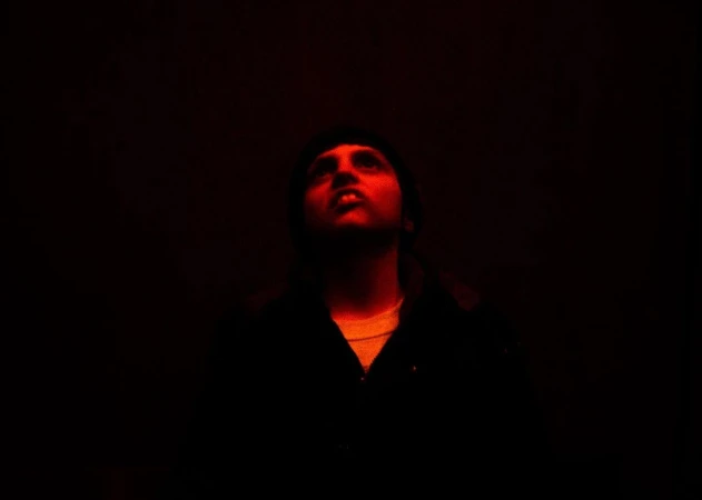 A man with dark hair looks upward in a dark room, illuminated by red lighting, wearing a dark jacket with a lighter shirt underneath.