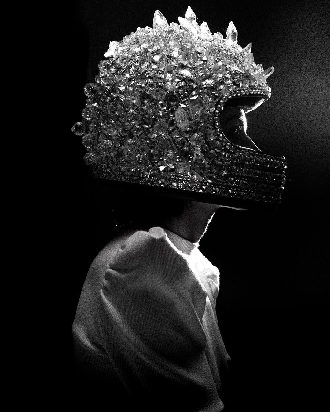 Profile shot of a person wearing a full-face motorcycle helmet entirely covered in crystals, against a dark background, in black and white.