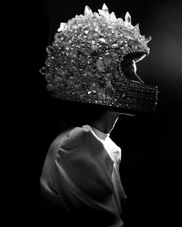 Profile shot of a person wearing a full-face motorcycle helmet entirely covered in crystals, against a dark background, in black and white.