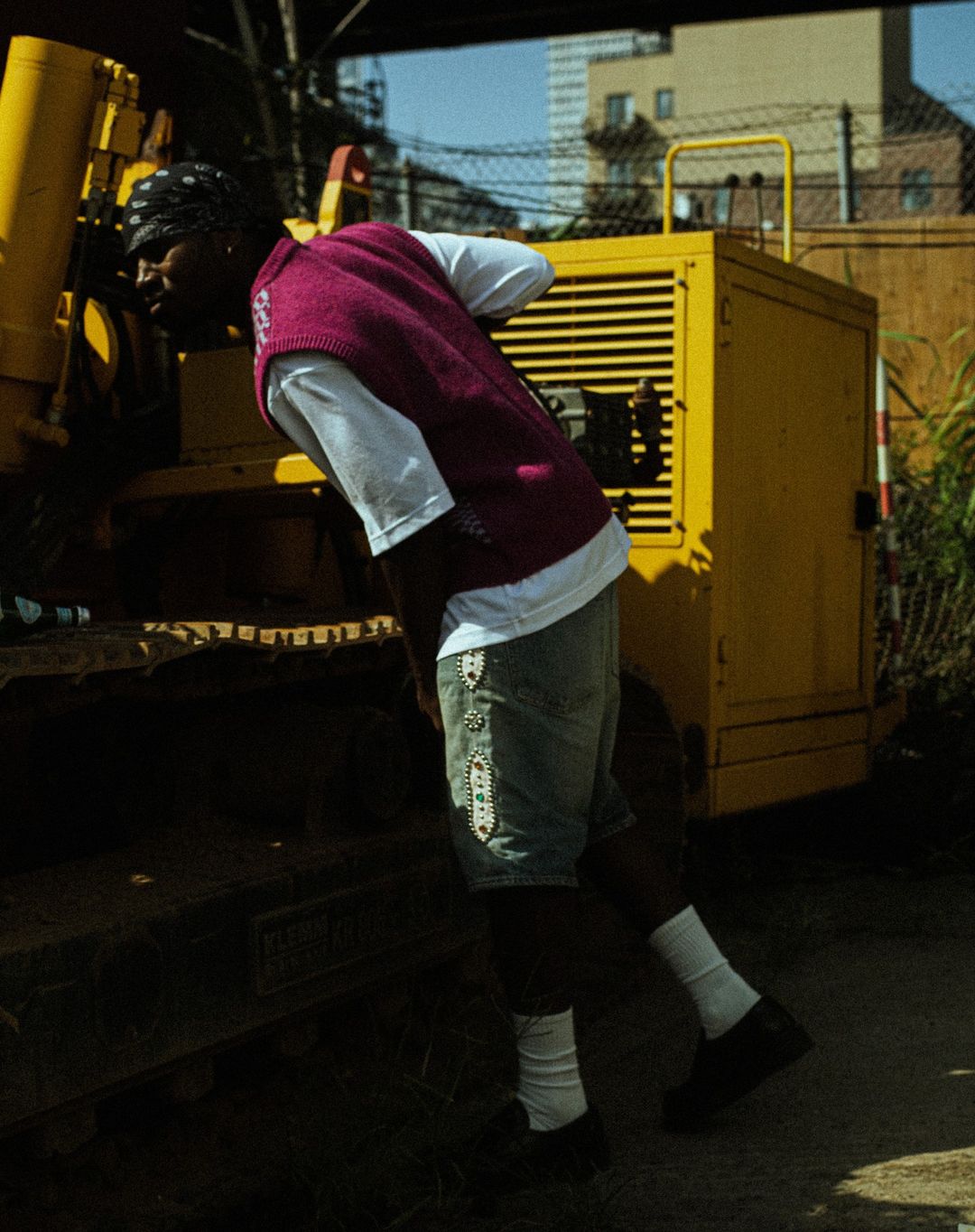 An African-American man is leaning on a yellow construction vehicle wearing a magenta vest and denim shorts outdoors on a sunny day.