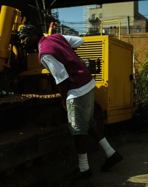 An African-American man is leaning on a yellow construction vehicle wearing a magenta vest and denim shorts outdoors on a sunny day.