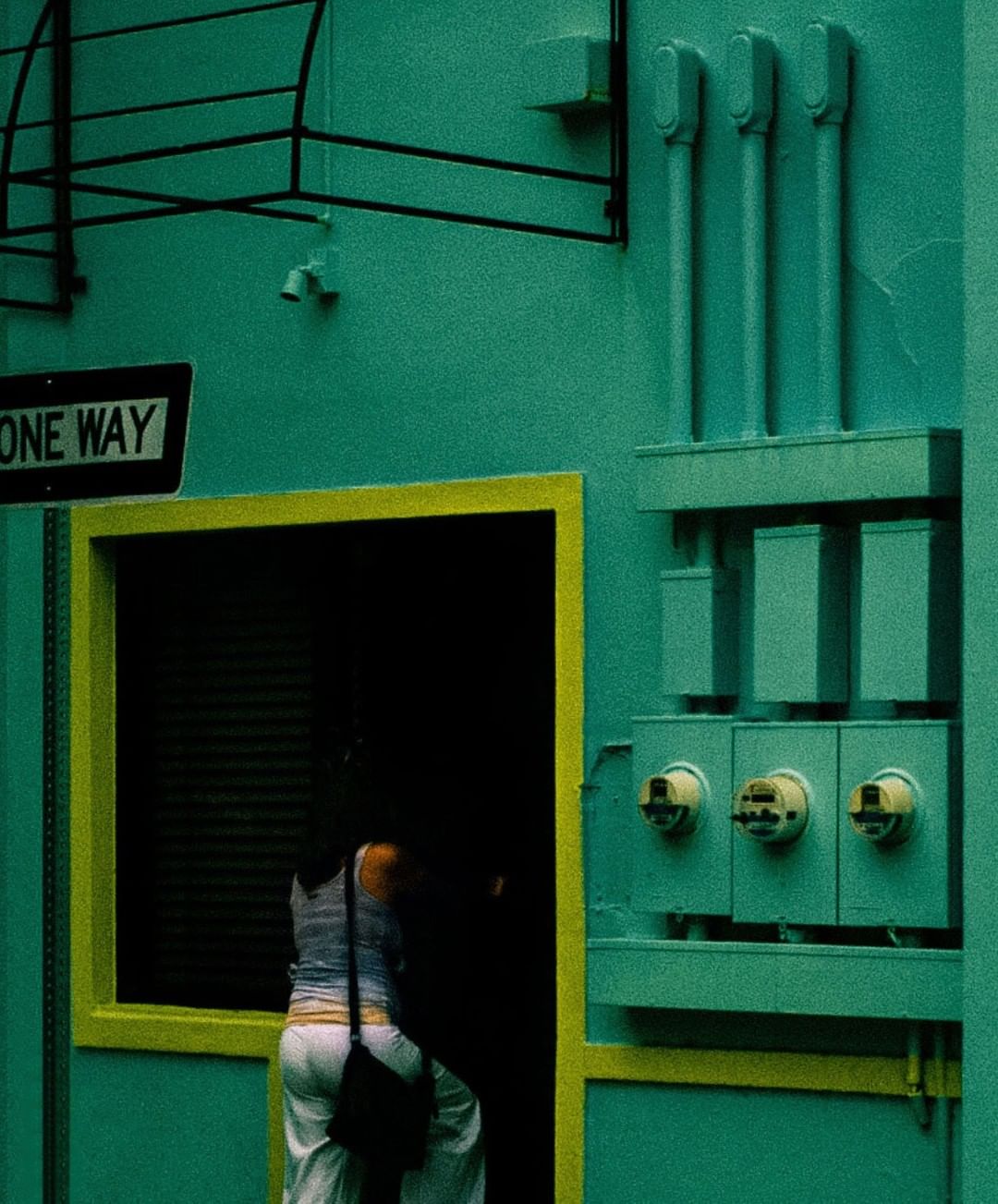 A woman walks through a yellow doorway of an industrial building painted in a vibrant green hue with utility boxes on the exterior.