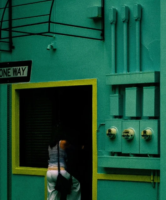 A woman walks through a yellow doorway of an industrial building painted in a vibrant green hue with utility boxes on the exterior.