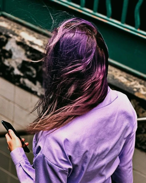 Back view of a woman with purple hair wearing a lilac shirt, holding a black mobile phone in an urban environment.