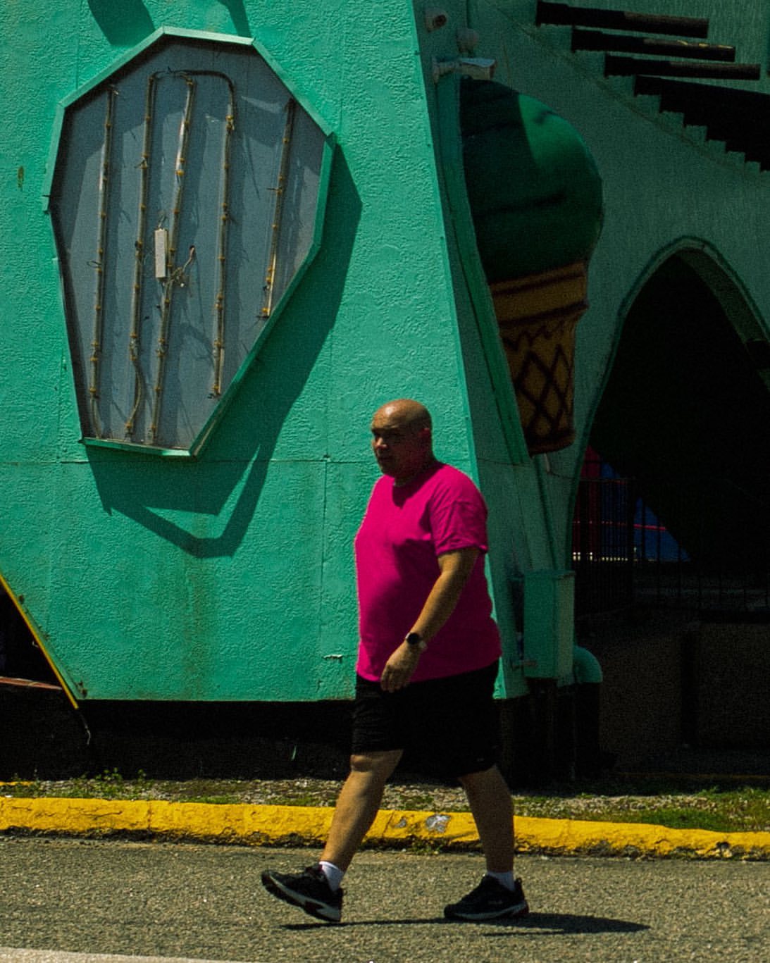 A man in a pink shirt walks on a sunny day in front of a turquoise building with a vintage sign and architectural details. 