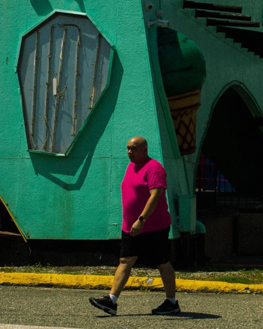 A man in a pink shirt walks on a sunny day in front of a turquoise building with a vintage sign and architectural details. 
