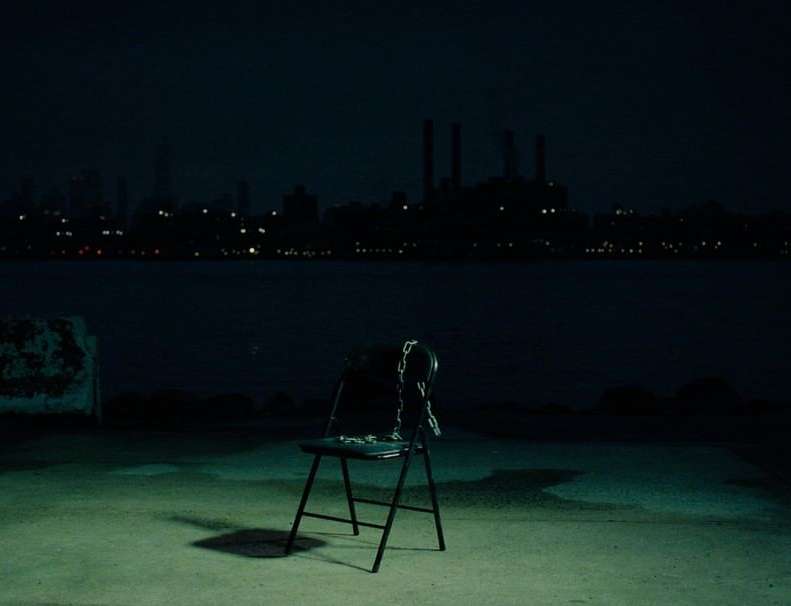 A single black chair sits on a concrete surface at night, overlooking a dark body of water and a distant cityscape with lights.