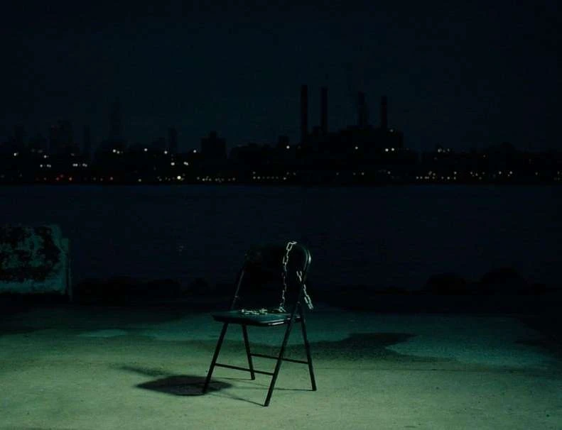 A single black chair sits on a concrete surface at night, overlooking a dark body of water and a distant cityscape with lights.