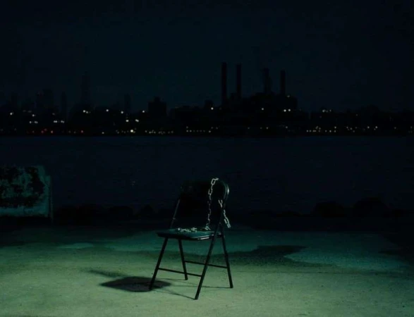 A single black chair sits on a concrete surface at night, overlooking a dark body of water and a distant cityscape with lights.