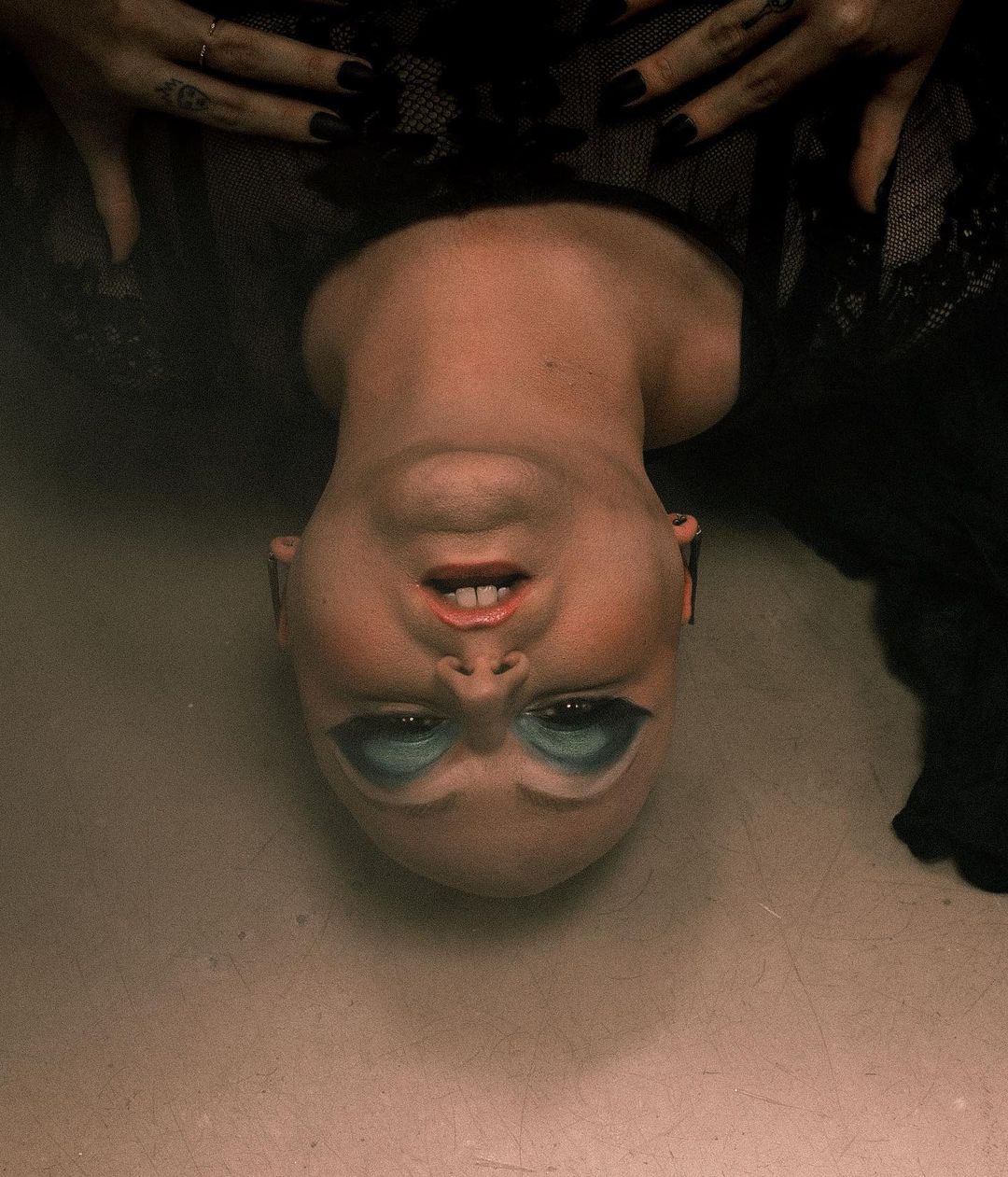 Close-up upside-down portrait of a bald female with blue eyeshadow and black lipstick, adorned in a black lace dress.