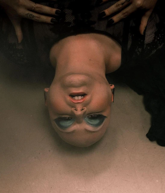Close-up upside-down portrait of a bald female with blue eyeshadow and black lipstick, adorned in a black lace dress.