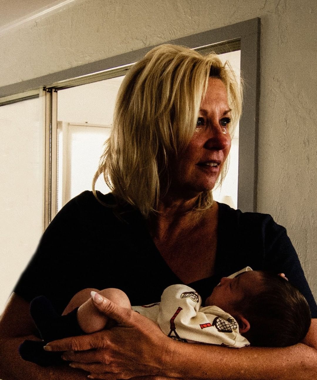 Indoor close-up of a crying woman with blond hair, holding a sleeping newborn baby in her arms, with a doorway visible behind them.