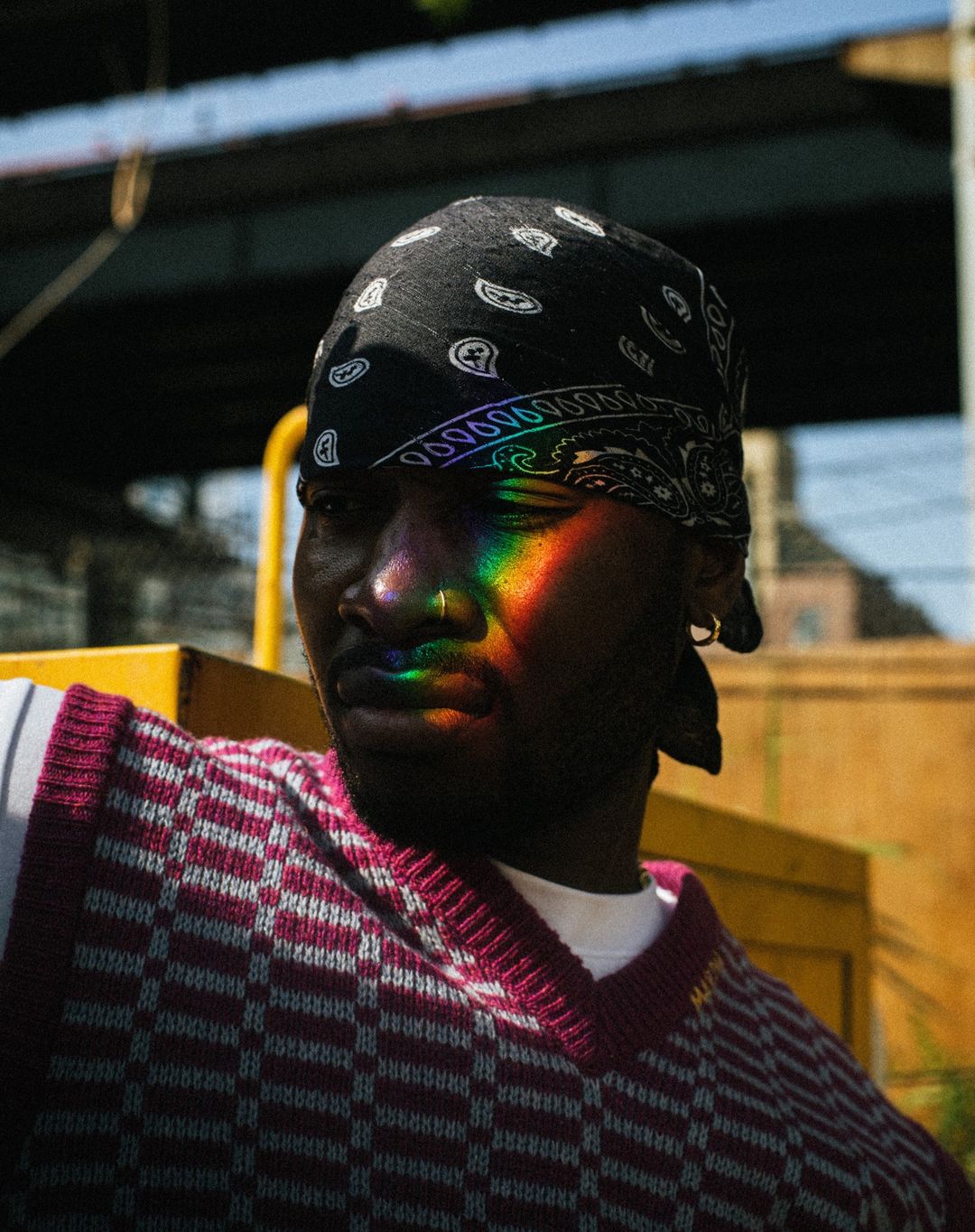 Portrait of a Black man wearing a bandana and vest, rainbow light on his face, captured outdoors under an overpass on a sunny day.