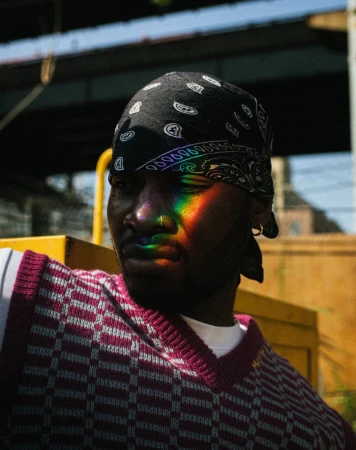 Portrait of a Black man wearing a bandana and vest, rainbow light on his face, captured outdoors under an overpass on a sunny day.