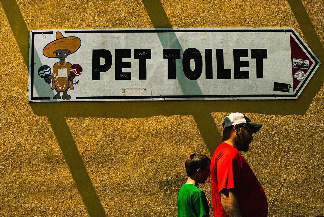 A father and son walking past a humorous sign on a yellow wall that reads, 'Pet Toilet,' with a cartoon dog illustration.