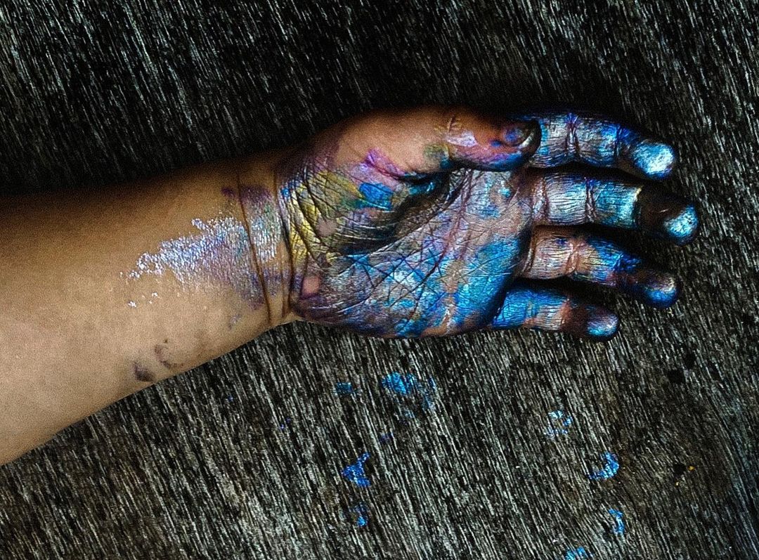Close-up of a hand covered in iridescent blue, gold, and violet paint against a dark gray textured background.