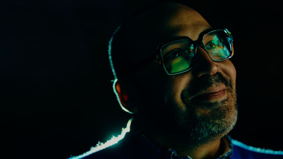 Close-up shot of an adult male with glasses smiling in dim lighting with a dark background. The man is wearing a collared shirt and jacket.