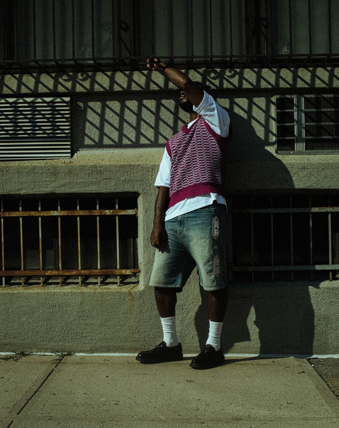 Adult male poses in front of a building wearing a white t-shirt, patterned vest, denim shorts, socks, and leather shoes in an urban setting.