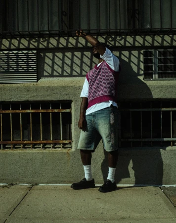 Adult male poses in front of a building wearing a white t-shirt, patterned vest, denim shorts, socks, and leather shoes in an urban setting.