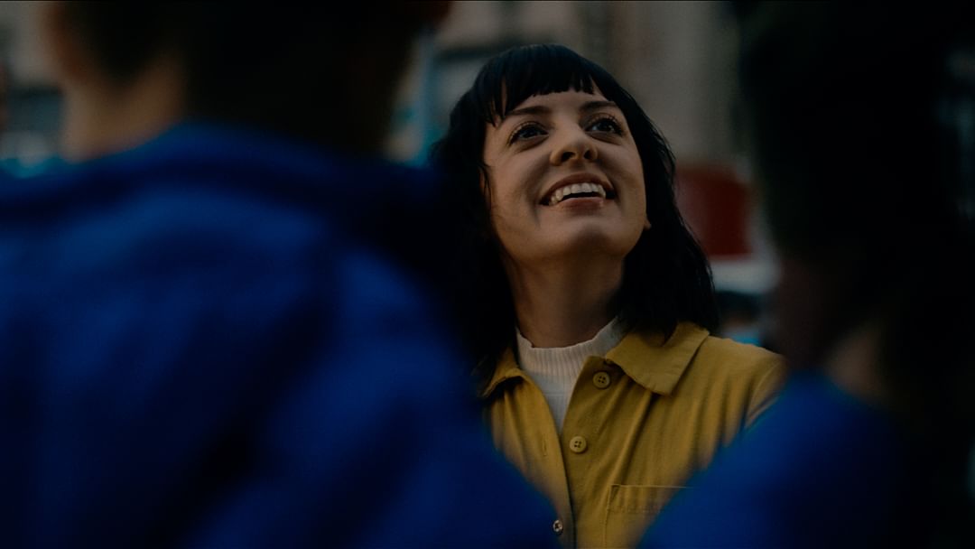 Smiling woman in yellow jacket surrounded by people, captured with a close-up shot, creating a warm and inviting scene.