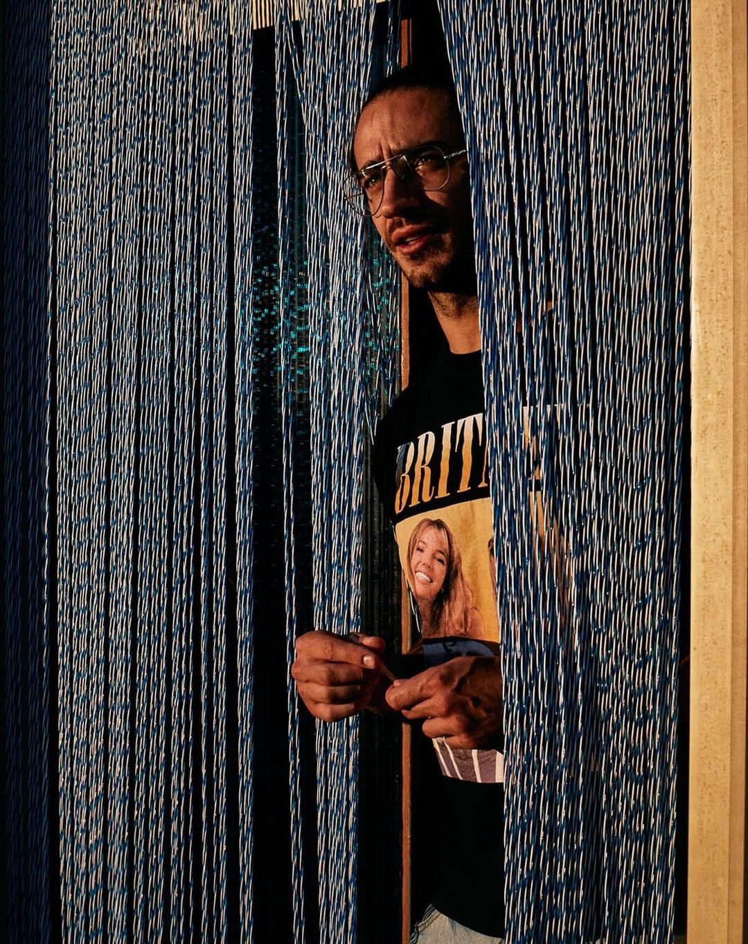 Man with glasses and a t-shirt with a blonde woman on it, standing behind hanging blue beaded curtains, looking off to the left.