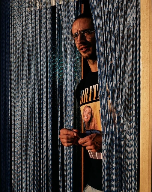 Man with glasses and a t-shirt with a blonde woman on it, standing behind hanging blue beaded curtains, looking off to the left.