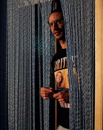 Man with glasses and a t-shirt with a blonde woman on it, standing behind hanging blue beaded curtains, looking off to the left.