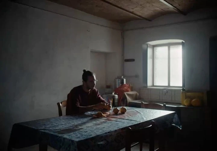 Man sitting at a table with fruit in a dimly lit interior, illuminated by window light, creating a contemplative and intimate scene.
