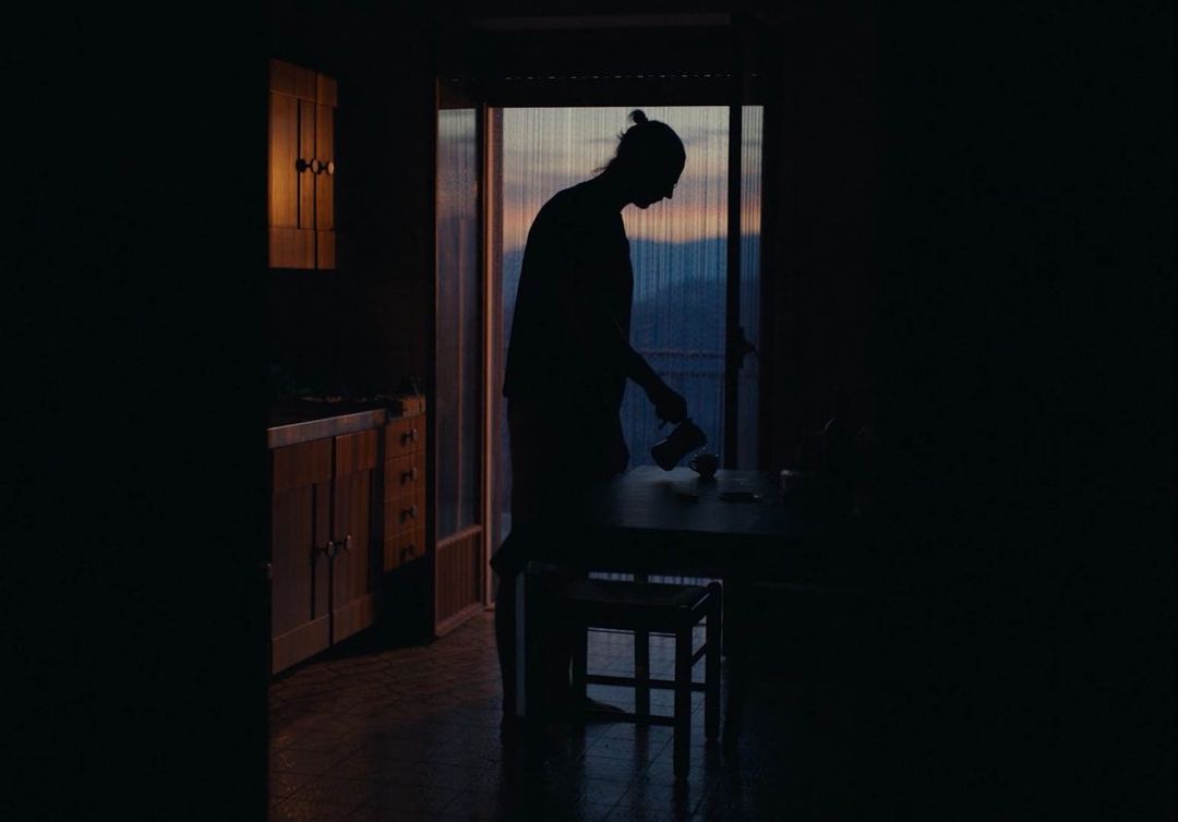 Silhouette of adult indoors near a table and chair, preparing coffee in an indoor environment at sunset with mountains in background.