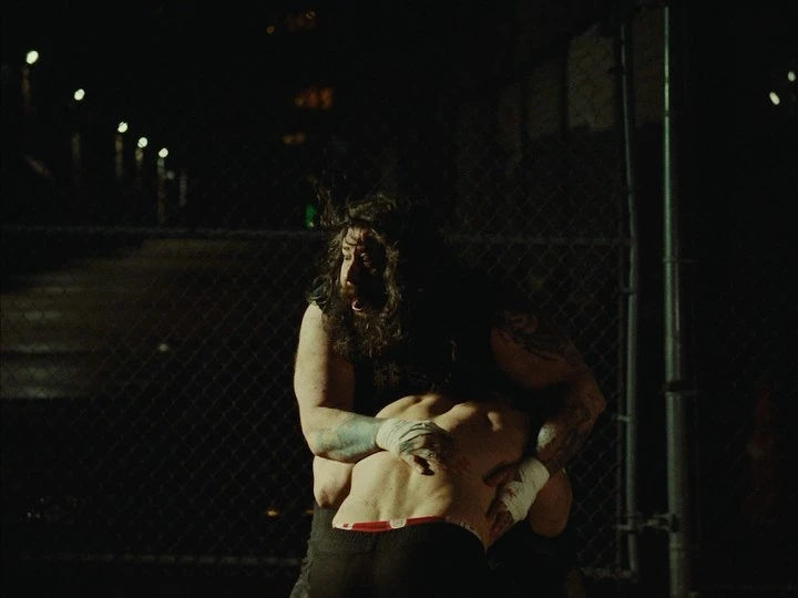 Two men wrestling outdoors at night. One has long hair and tattoos. The other man is lifted in a fireman carry. Dark, gritty.