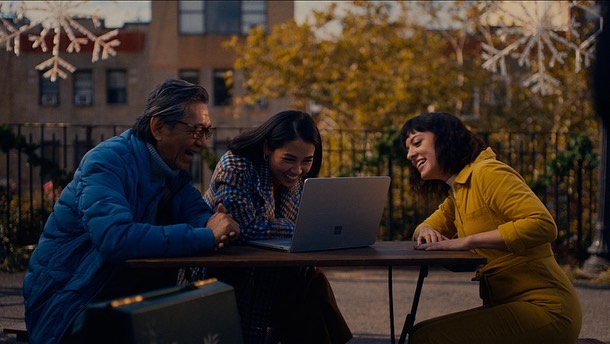 Three people smiling and looking at a laptop outdoors near bare trees and decorations, wearing casual clothing.