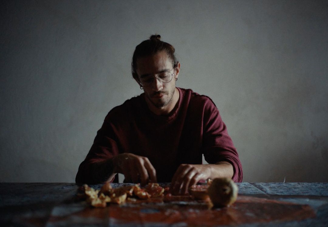 Man with glasses and hair in a bun sits indoors, sorting orange peels on a table with a patterned tablecloth in low light.