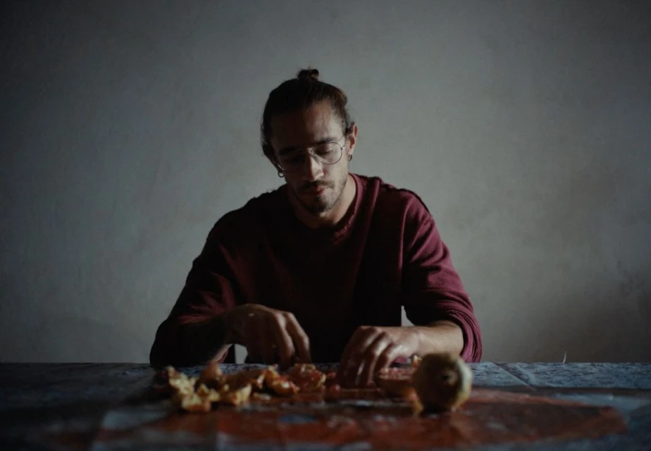 Man with glasses and hair in a bun sits indoors, sorting orange peels on a table with a patterned tablecloth in low light.