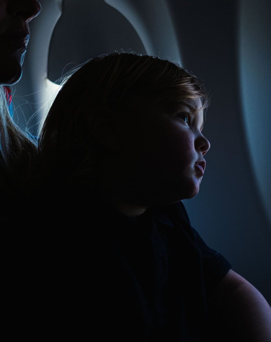 Child looks out the window of an airplane in a dramatically lit close-up, accompanied by an adult. The window provides a soft blue background.