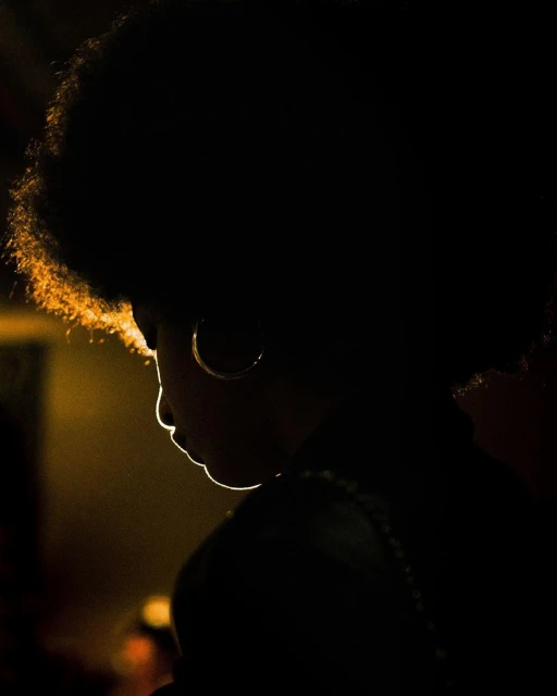 Silhouette of a woman with an afro hairstyle and hoop earrings in a dimly lit environment with warm backlight illuminating her features.