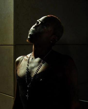 Portrait of a black man with short red hair wearing a black top and silver cross necklace, captured in dramatic low-key lighting.