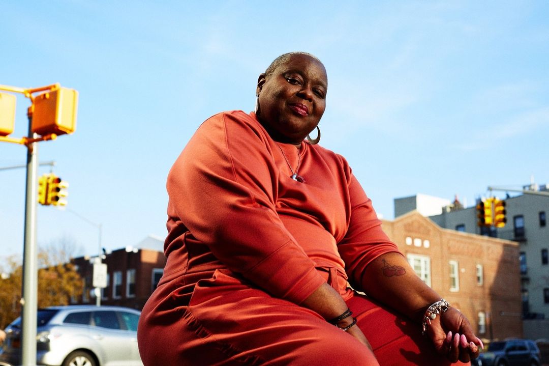 Portrait of an adult woman in orange clothing, smiling, outdoors in an urban setting, with buildings and traffic lights visible.