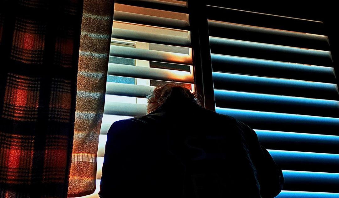 Back view of a person standing by a window with blinds, looking out; curtain partially obscures view. Dramatic lighting creates strong shadows.