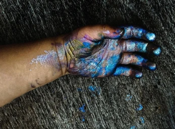 Close-up of a human hand and forearm resting on a dark wood grain surface, covered in iridescent blue and purple metallic paint.