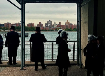 Several individuals in traditional black coats and hats stand along a waterfront railing overlooking a city skyline across a river.